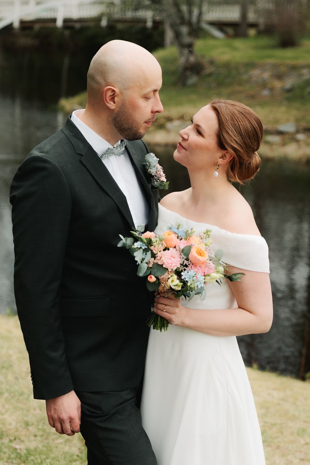 Wedding couple embraces on grassy lawn beside a tranquil pond during their spring celebration.