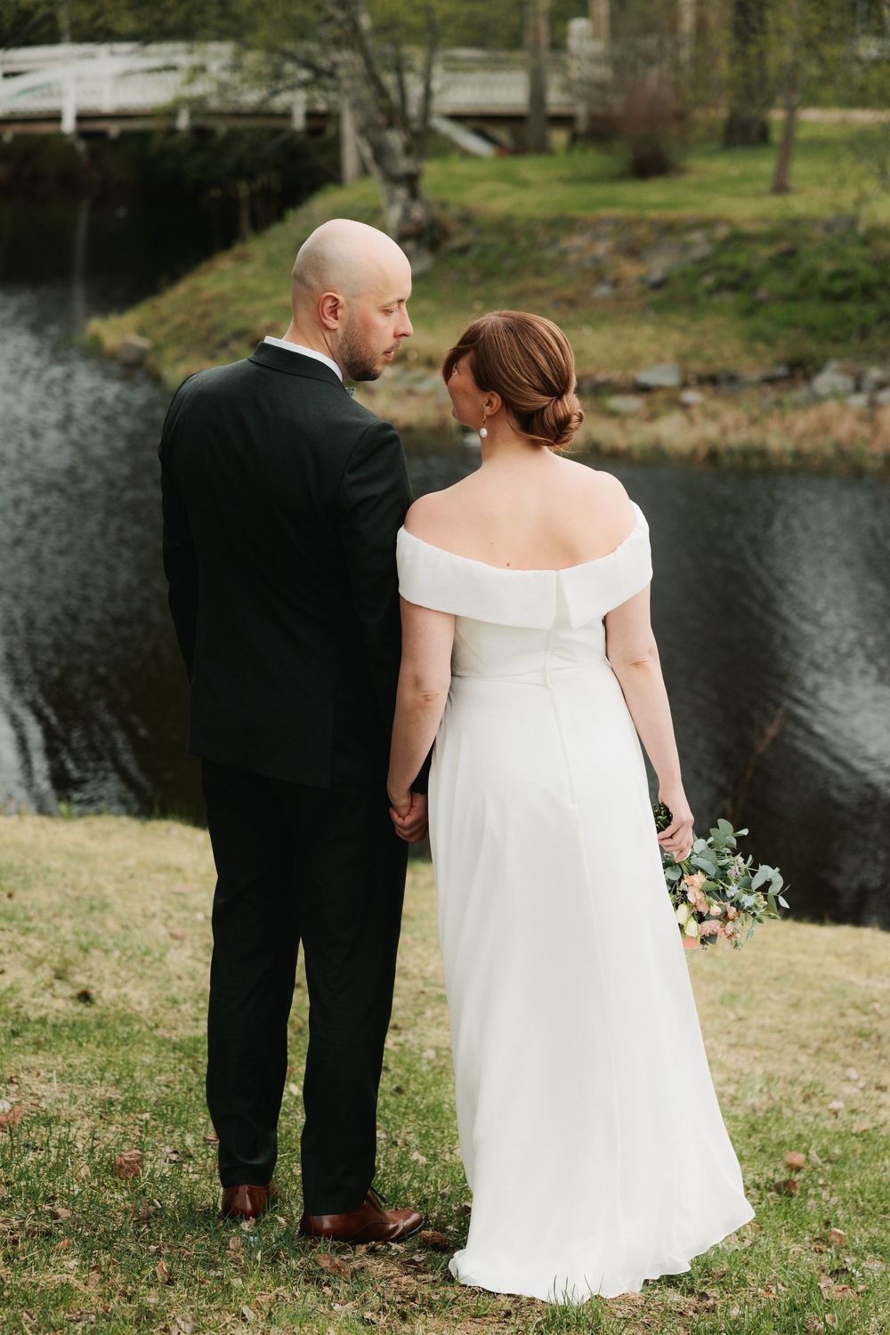 A couple in wedding attire stands by a serene lake, with the bride wearing an elegant off-shoulder white gown.