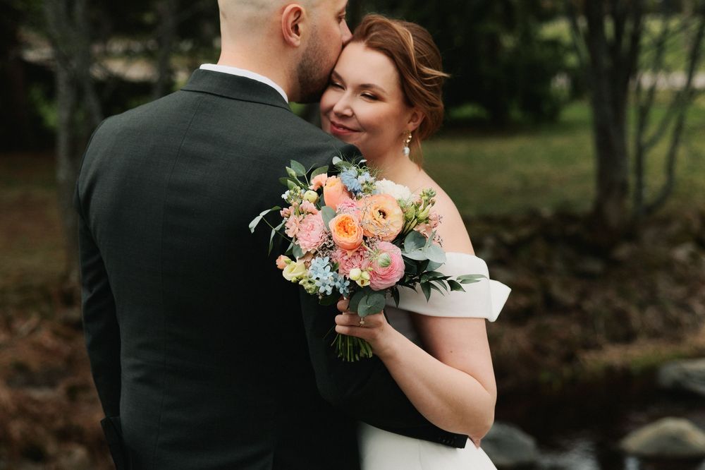 A romantic wedding couple embraces in a lush garden setting with soft peach and white floral bouquet details.