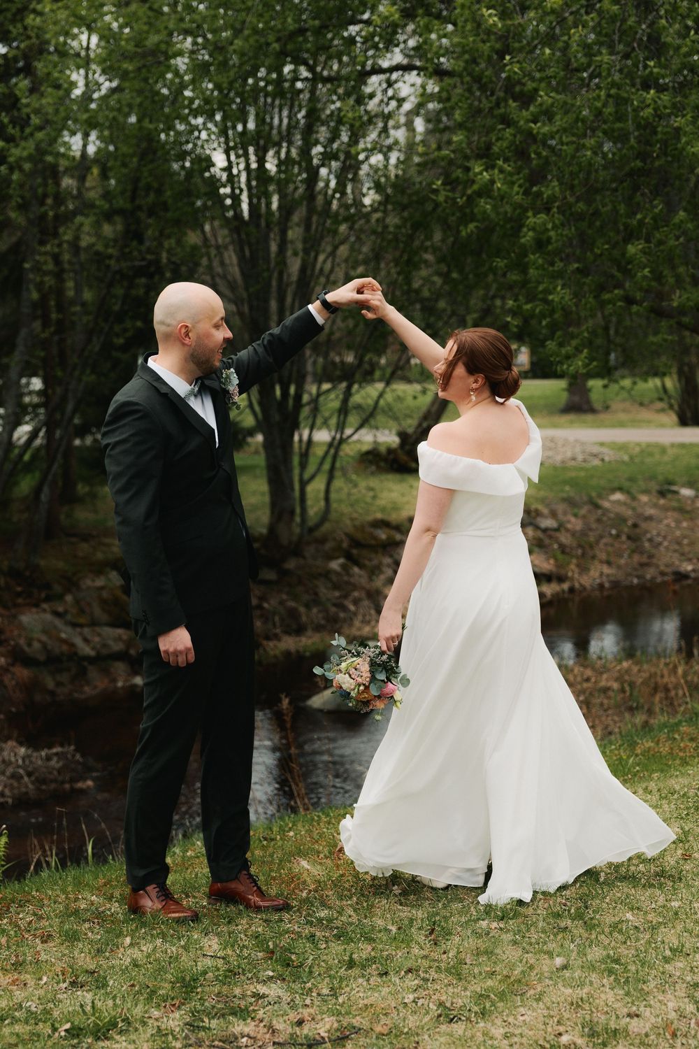 Newlyweds share a playful moment by a pond in a lush outdoor setting with the bride twirling her white dress.