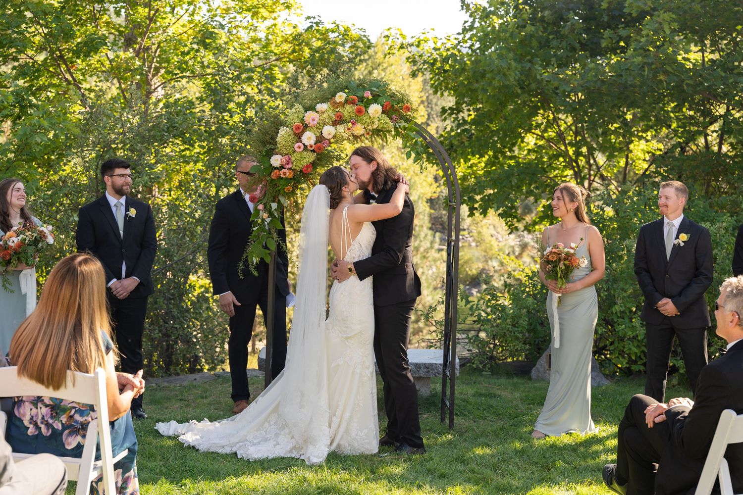 A romantic outdoor wedding ceremony takes place under lush green trees in a garden setting.