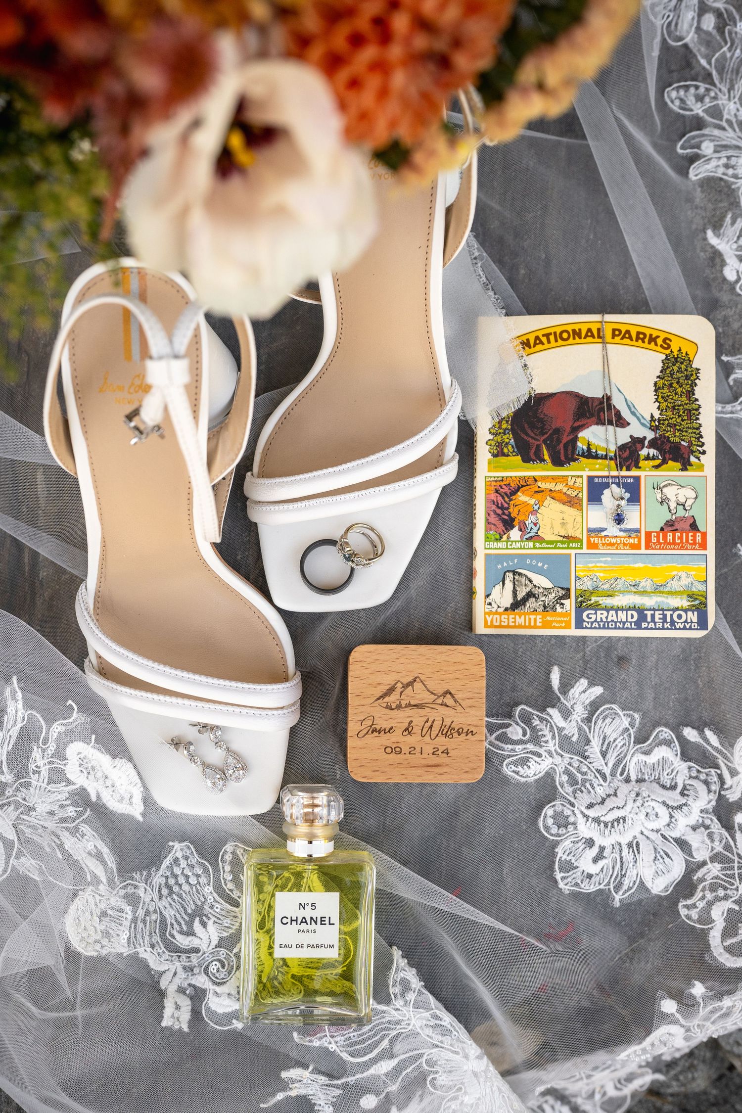Wedding details flatlay with white heels, perfume bottle, and vintage postcard on lace fabric.