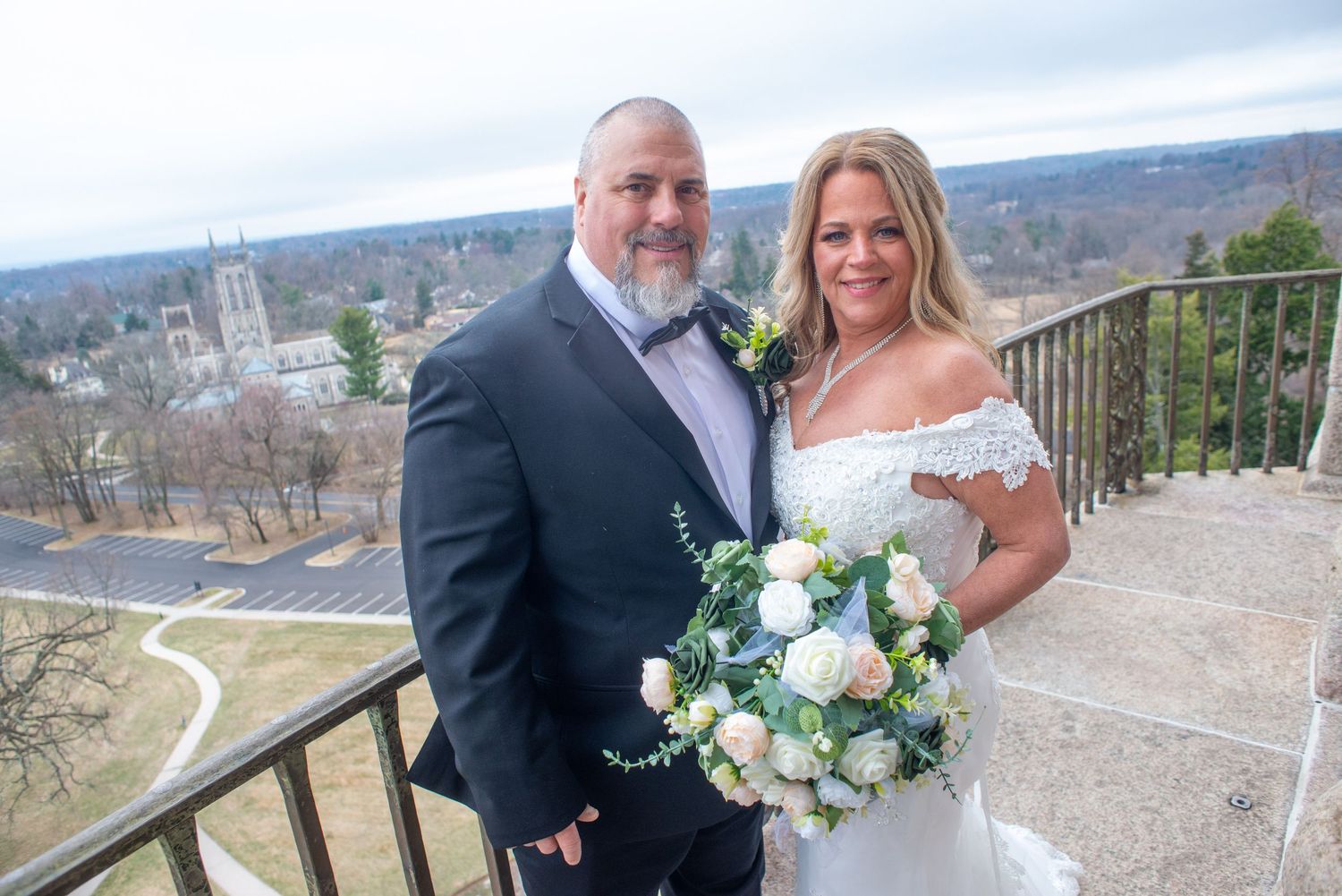 A couple poses on a scenic overlook with a white floral bouquet and church steeple visible in the background.