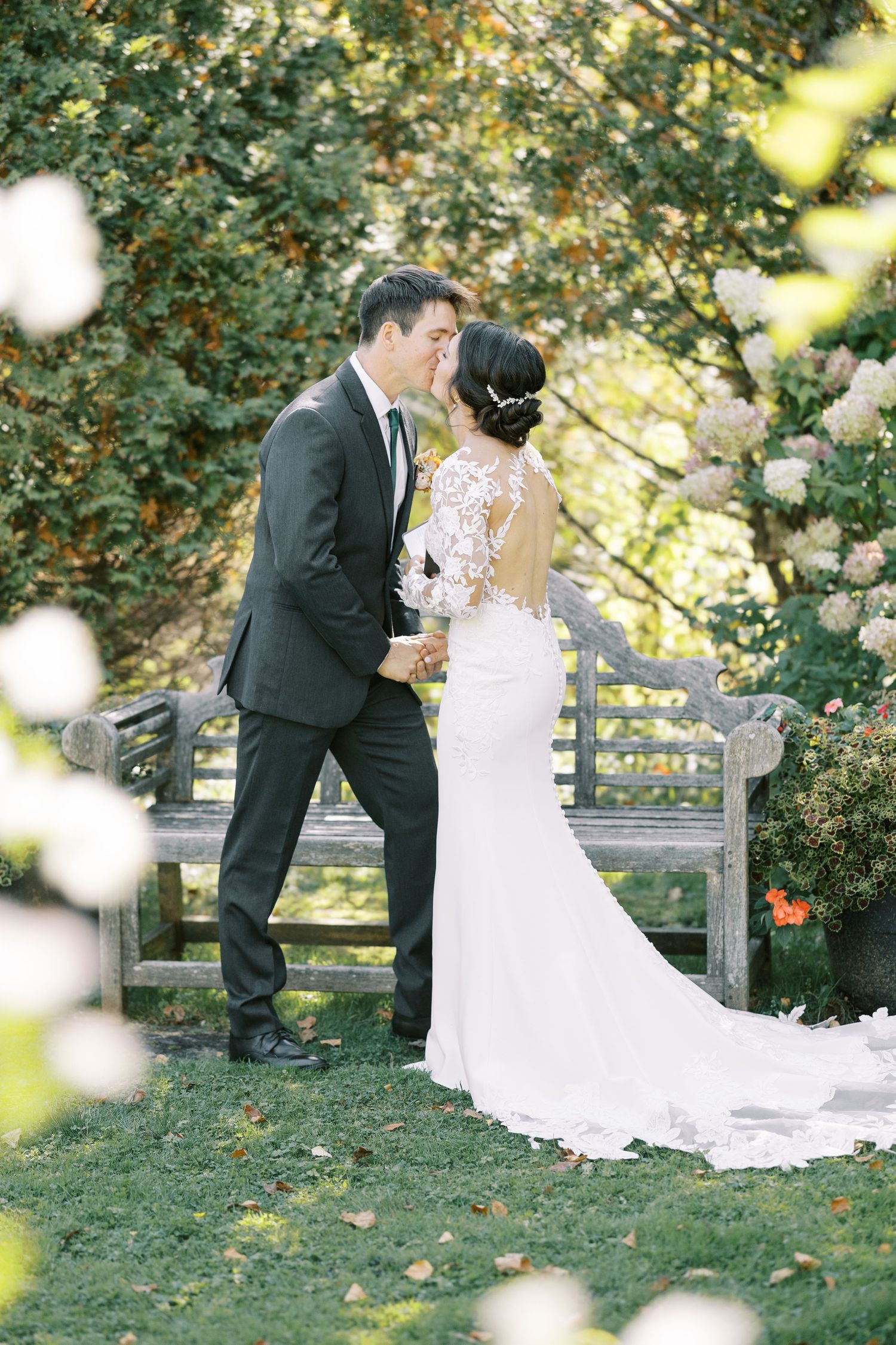 A romantic wedding couple shares intimate moments on a garden bench surrounded by lush greenery and soft bokeh lighting.