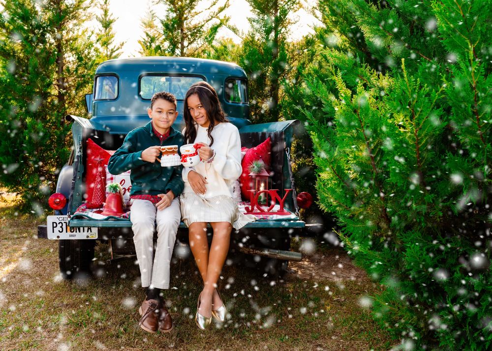 Family poses by vintage blue car with evergreen trees and falling snow creating a festive winter scene.