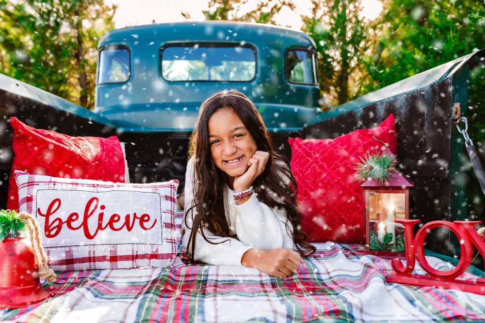 A festive winter scene with a red truck, snow, and holiday decorations in a rustic outdoor setting.