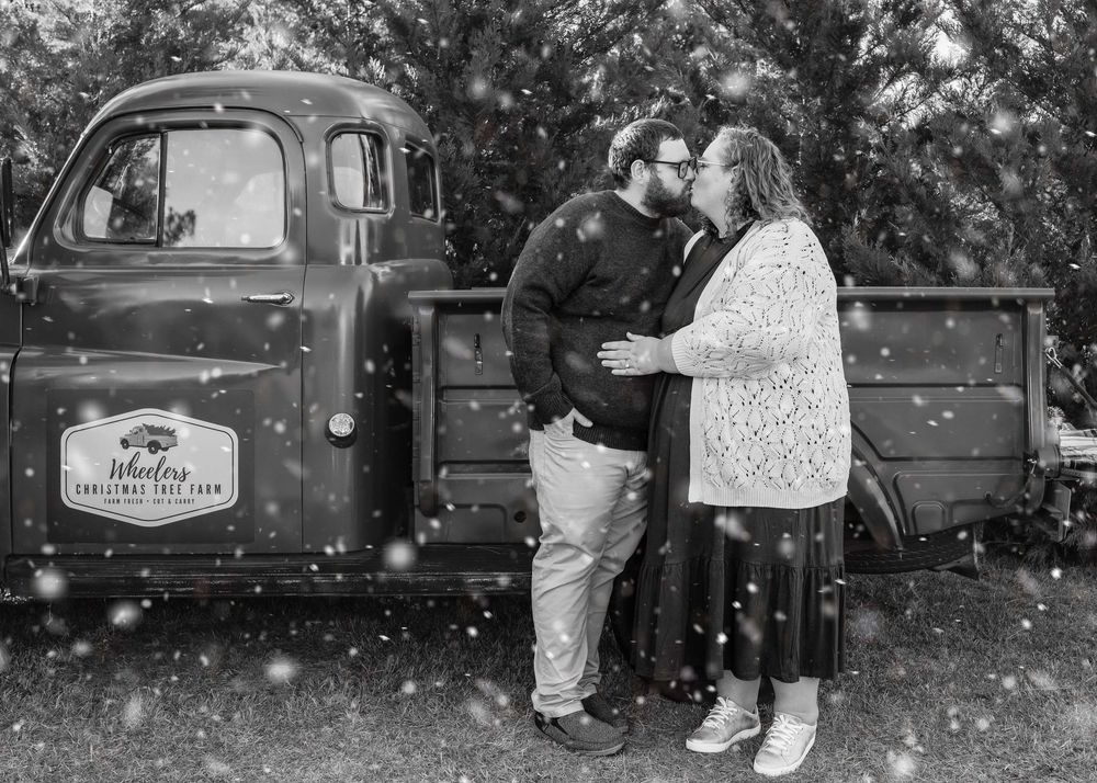 A couple stands in front of a vintage pickup truck with falling snow in a romantic black and white scene.
