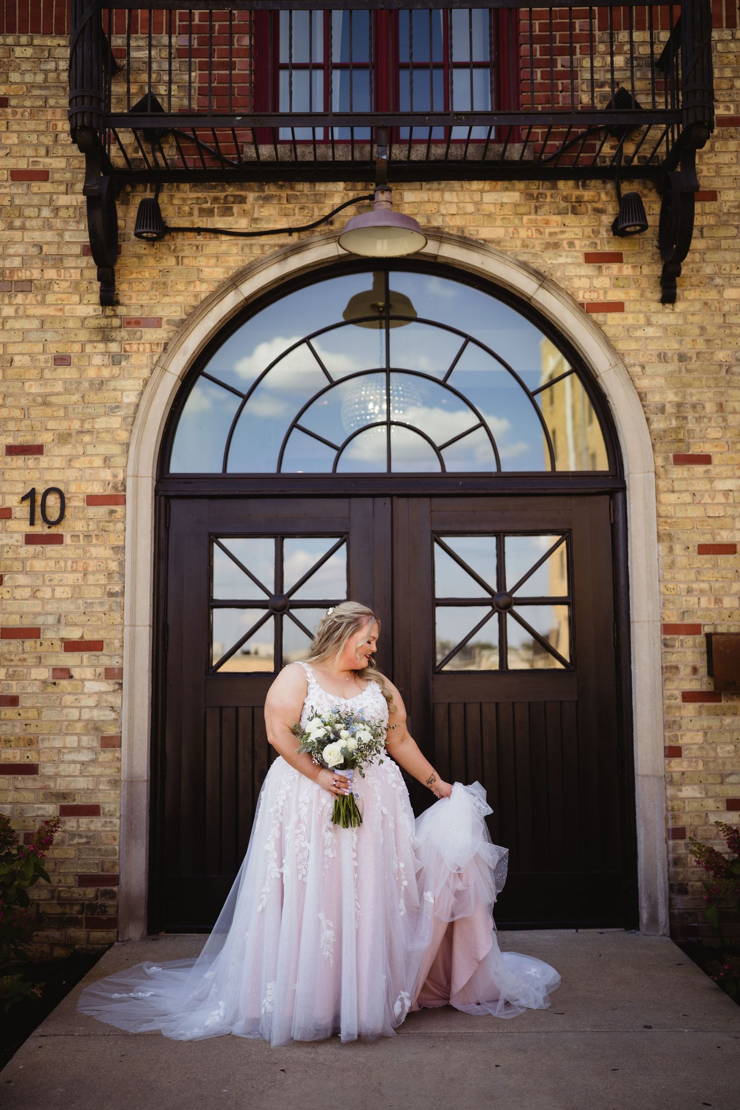 A bride in a flowing pink and white gown poses elegantly in front of a rustic brick building with arched doorways.