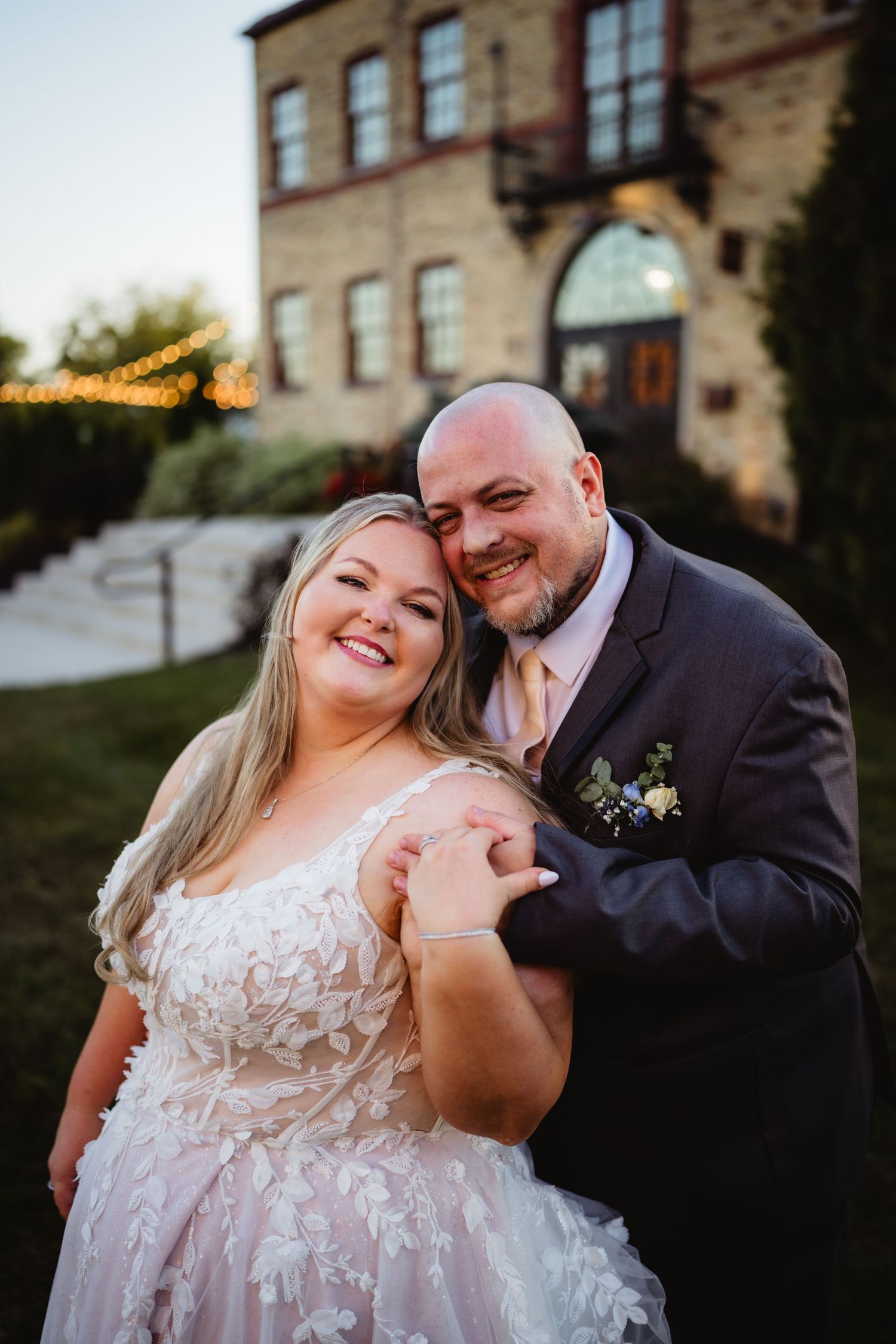 A happy couple shares intimate moments in formal wedding attire against a stone building backdrop at sunset.