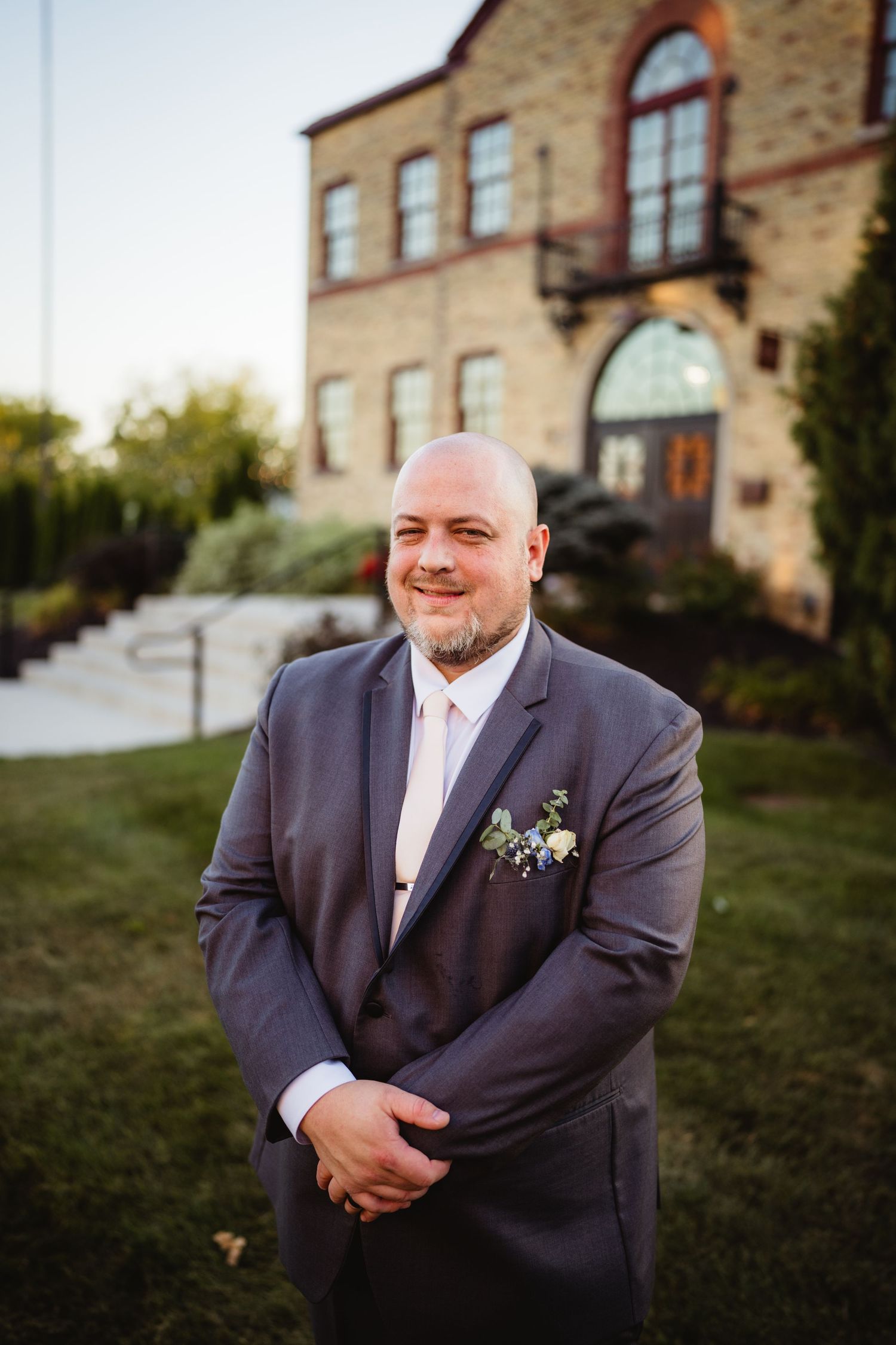 A groom in a dark suit and white boutonniere poses outdoors against a stone building backdrop during golden hour.