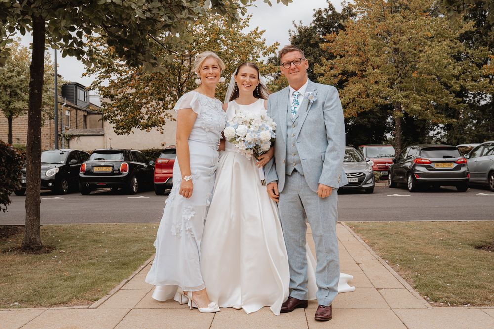 Bride in white wedding gown poses with parents outdoors on sunny wedding day.