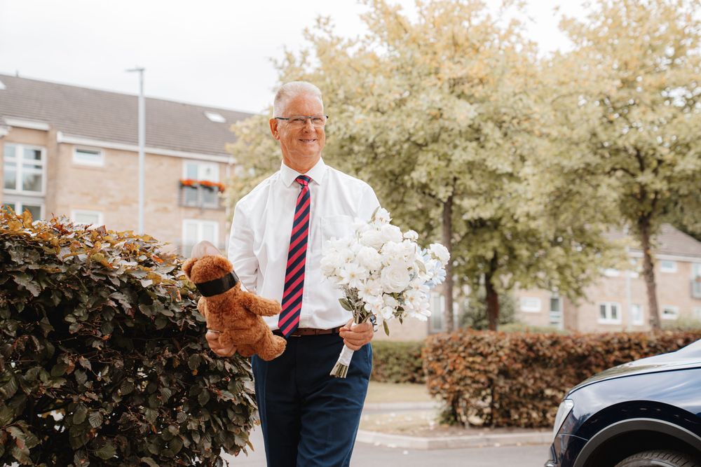 Smiling older gentleman carrying white bridal bouquet and stuffed toy outside on wedding day.
