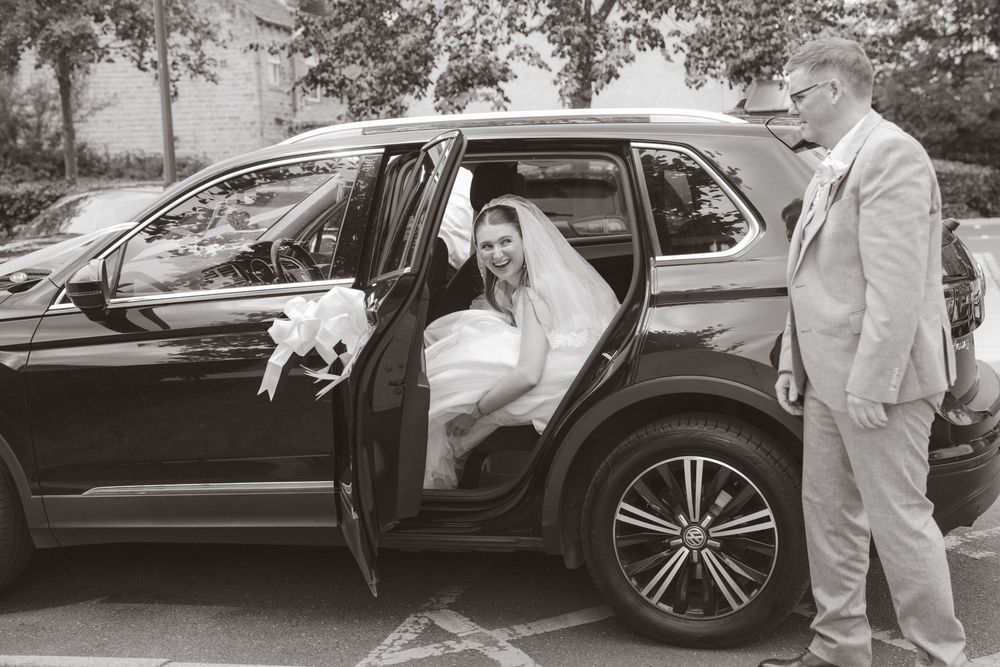 Bride in white dress exits black SUV wedding car assisted by man in light suit.