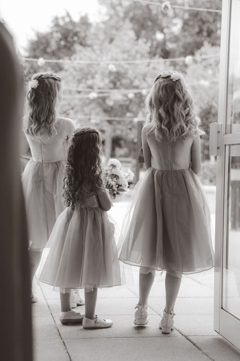 Three flower girls in tulle dresses with curled hair stand at venue entrance.