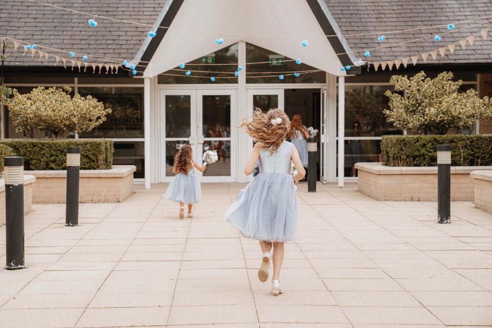 Two flower girls in matching light blue dresses running toward decorated wedding venue entrance.