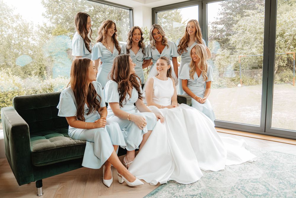 Bride in white gown seated with seven bridesmaids in matching light blue dresses by large windows.