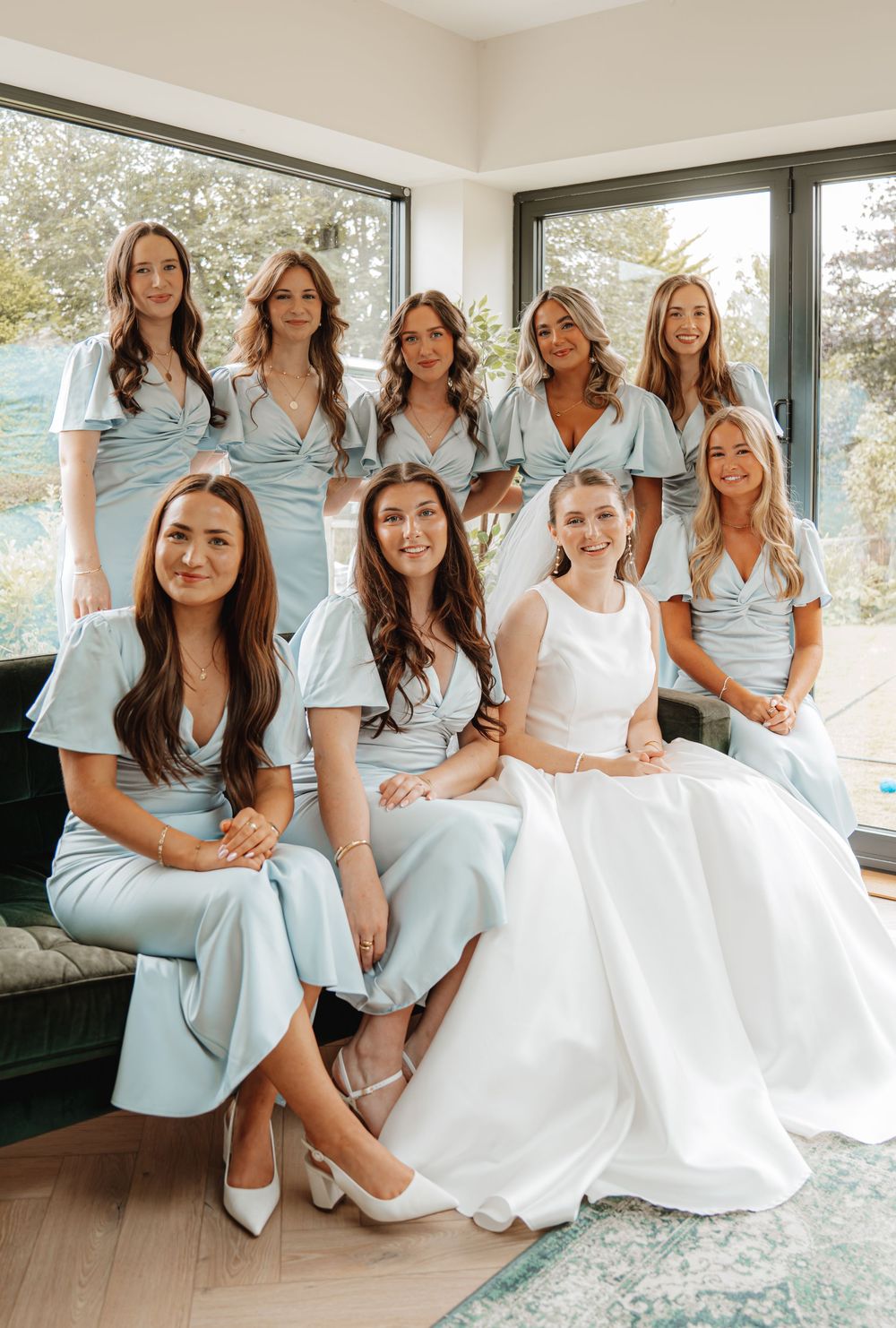 Bride posing with eight bridesmaids wearing coordinating light blue midi dresses in a bright modern room.