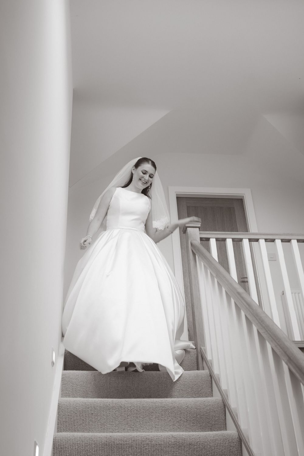 Elegant bride in full white ballgown and veil gracefully descending a staircase in black and white photo.