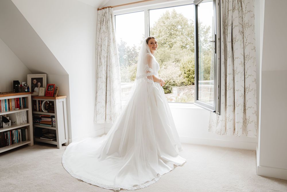 Bride in stunning white ballgown with long cathedral veil standing by a sunlit window in a bright room.