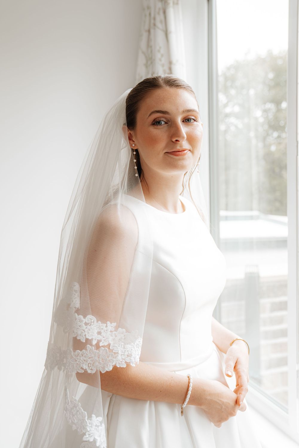 Beautiful bride in white lace-sleeved wedding dress and veil smiling softly while standing near a bright window.
