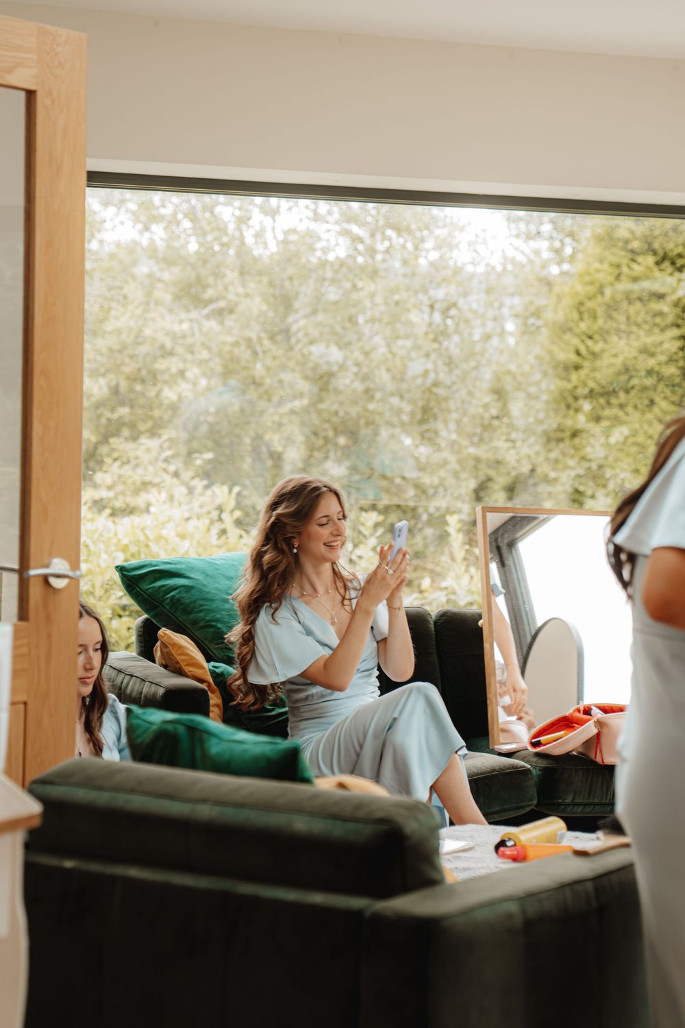 Bridesmaid in light blue dress relaxing on sofa using phone during wedding morning preparations.