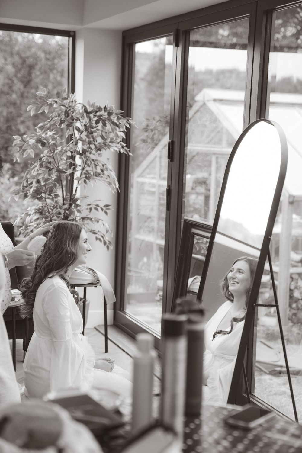 Bride in white robe smiling at her reflection in a full-length mirror during wedding preparations.