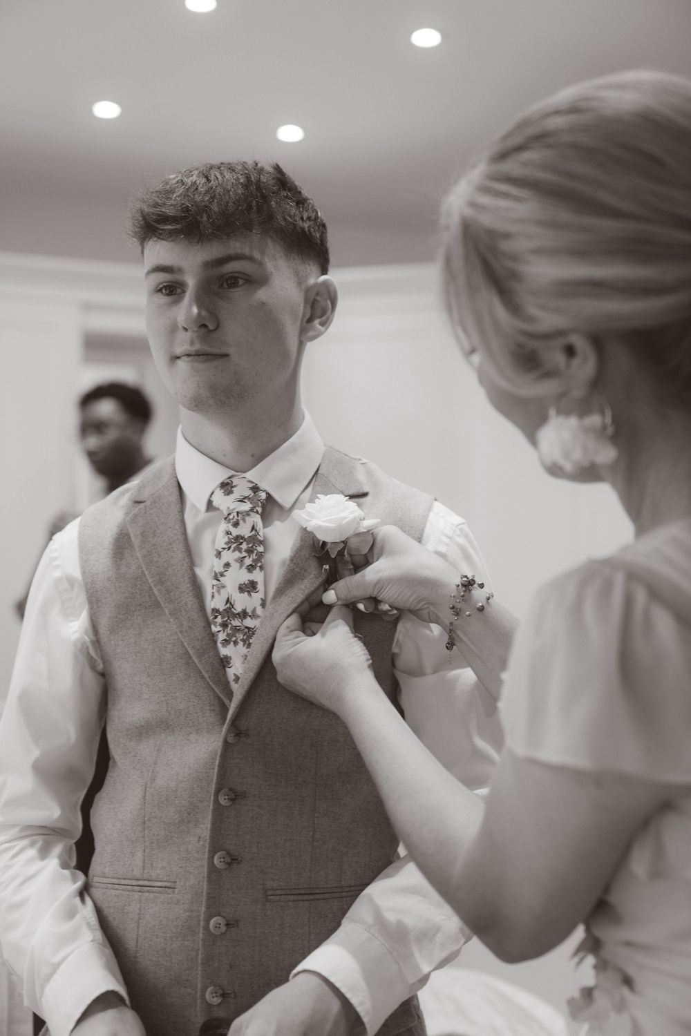 Woman pinning a white boutonniere onto a young mans grey wedding vest and floral tie.