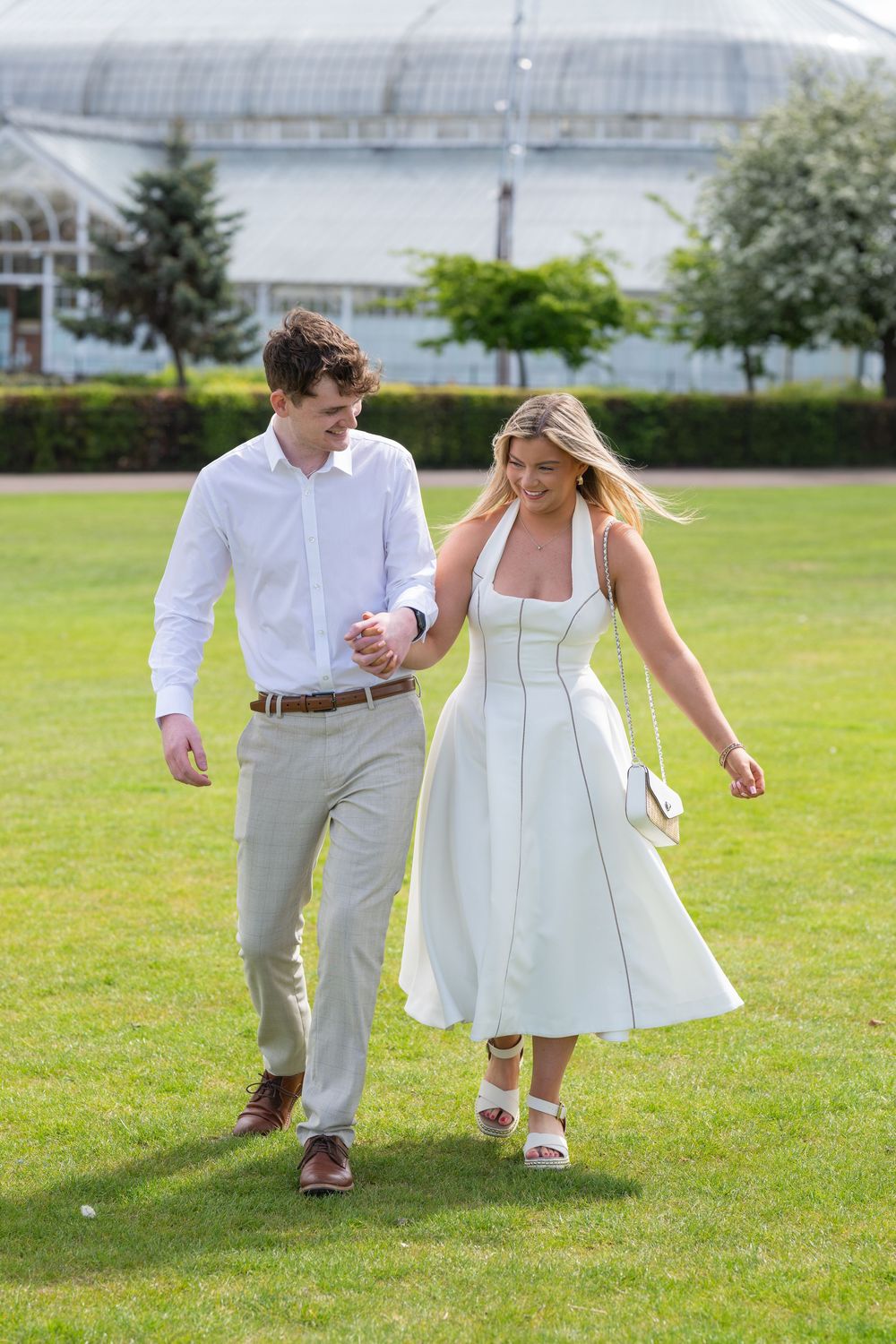 A couple strolls hand in hand across a lush green lawn in summer wedding attire.