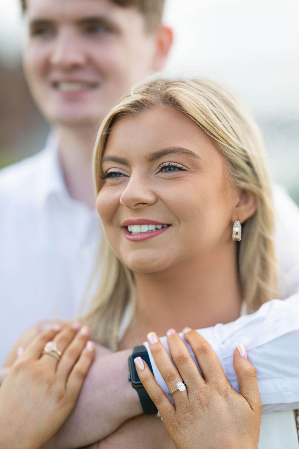 A close-up portrait showing a bright smile and sparkling engagement ring against a light background.