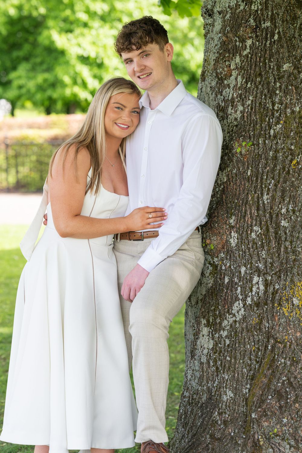 Couple in white clothing poses for romantic engagement photos against a large tree in a sunny outdoor setting.
