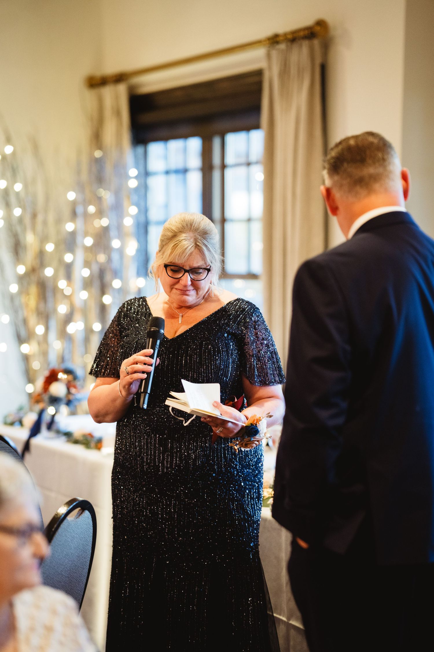 Woman in sparkly black dress reads from paper at formal event with twinkle lights in background.