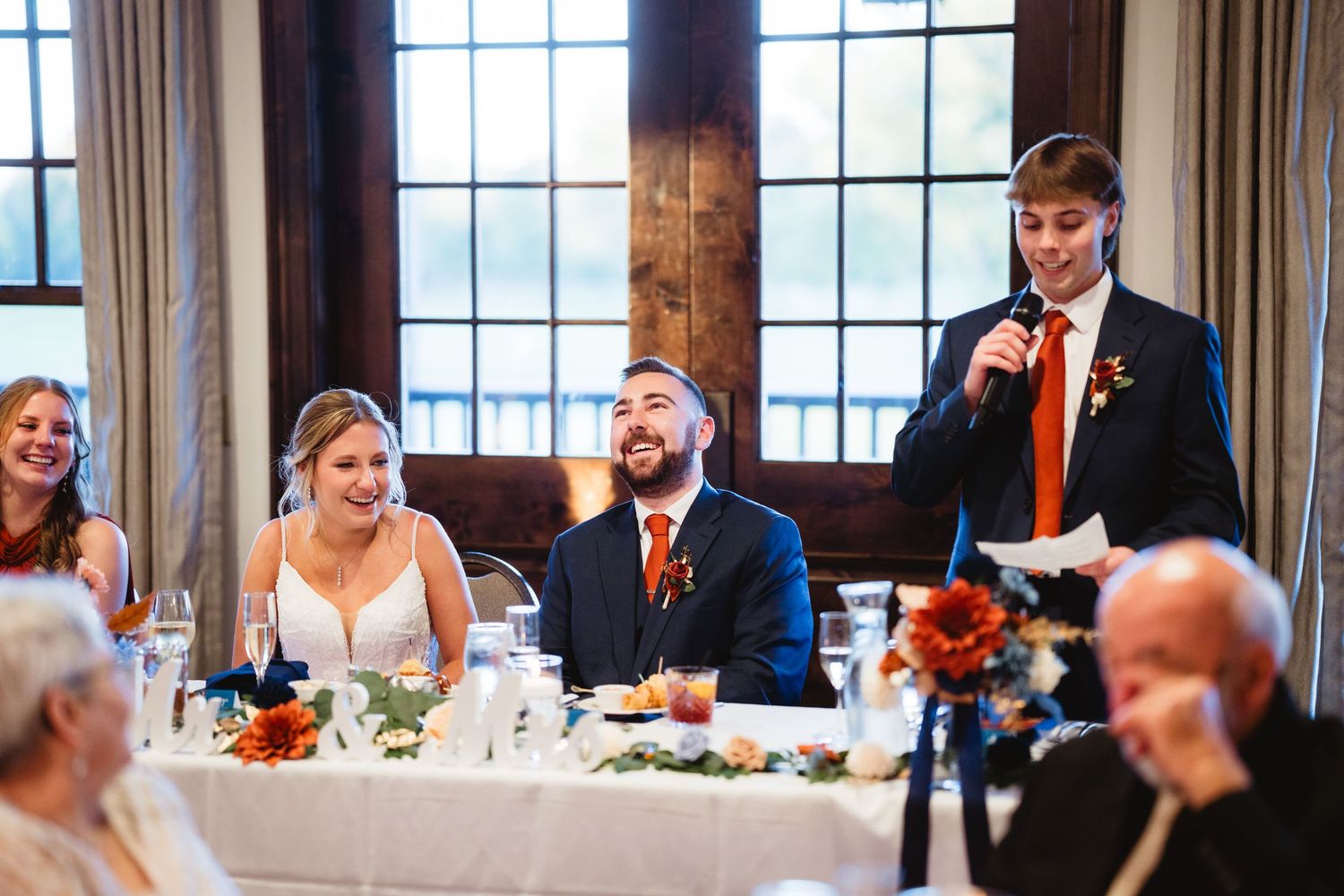 Wedding guests seated at long tables in an elegant dining room share joyful laughter during a reception celebration.