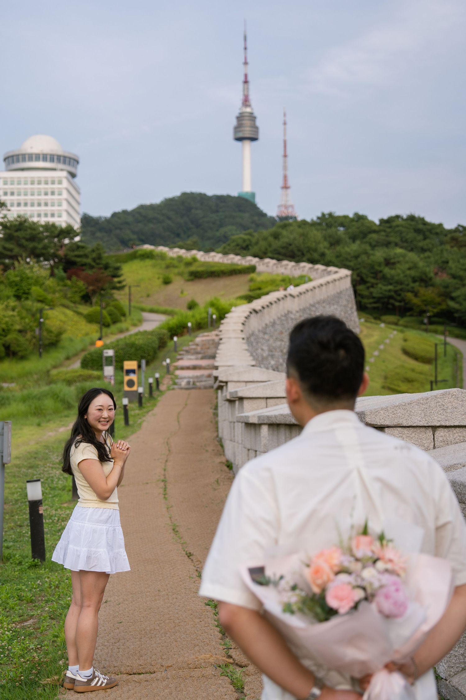 A scenic proposal moment unfolds on a hilltop path with Seoul Tower visible in the background.