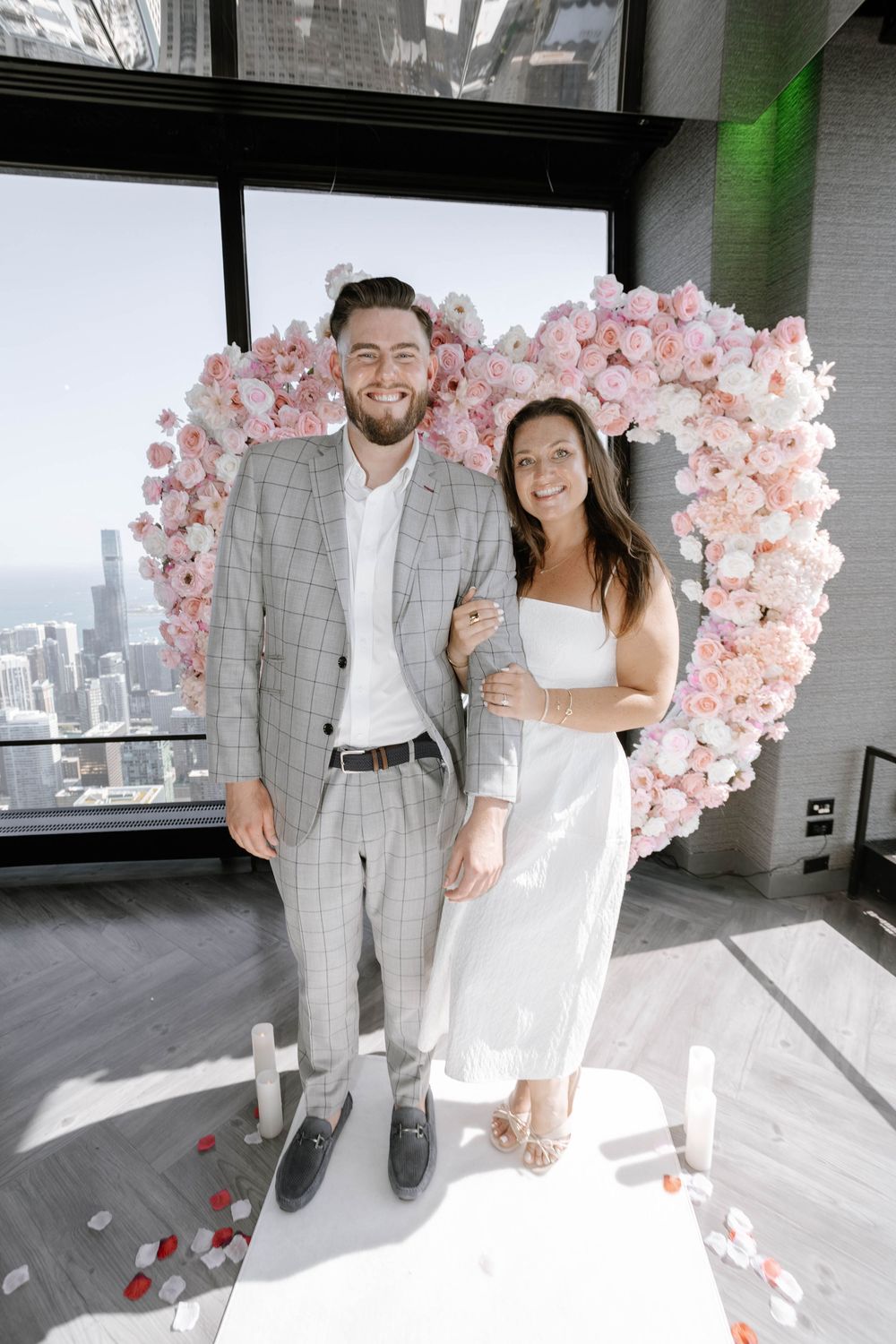 A couple poses together in front of a pink floral arch in a modern indoor space with city views.