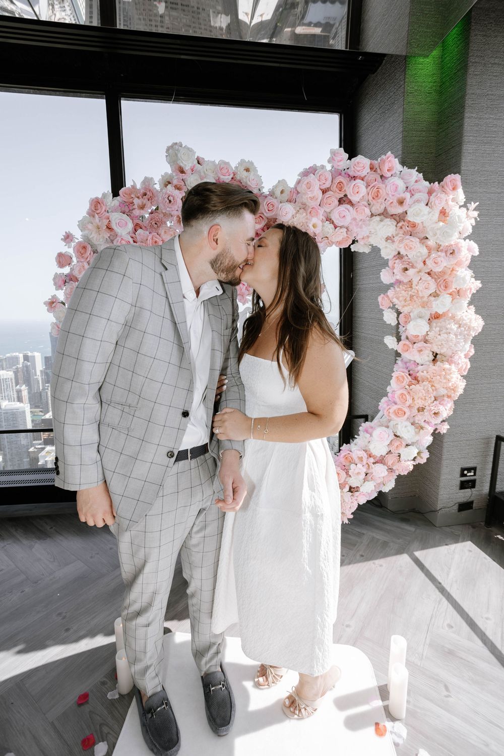 A romantic wedding scene with a delicate pink floral arch and a couple in white and gray attire in an indoor setting.