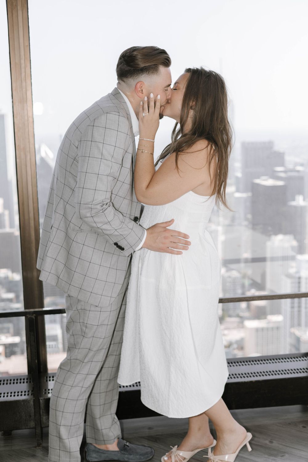 A romantic couple in grey and white attire share an intimate moment by large windows overlooking a city skyline.