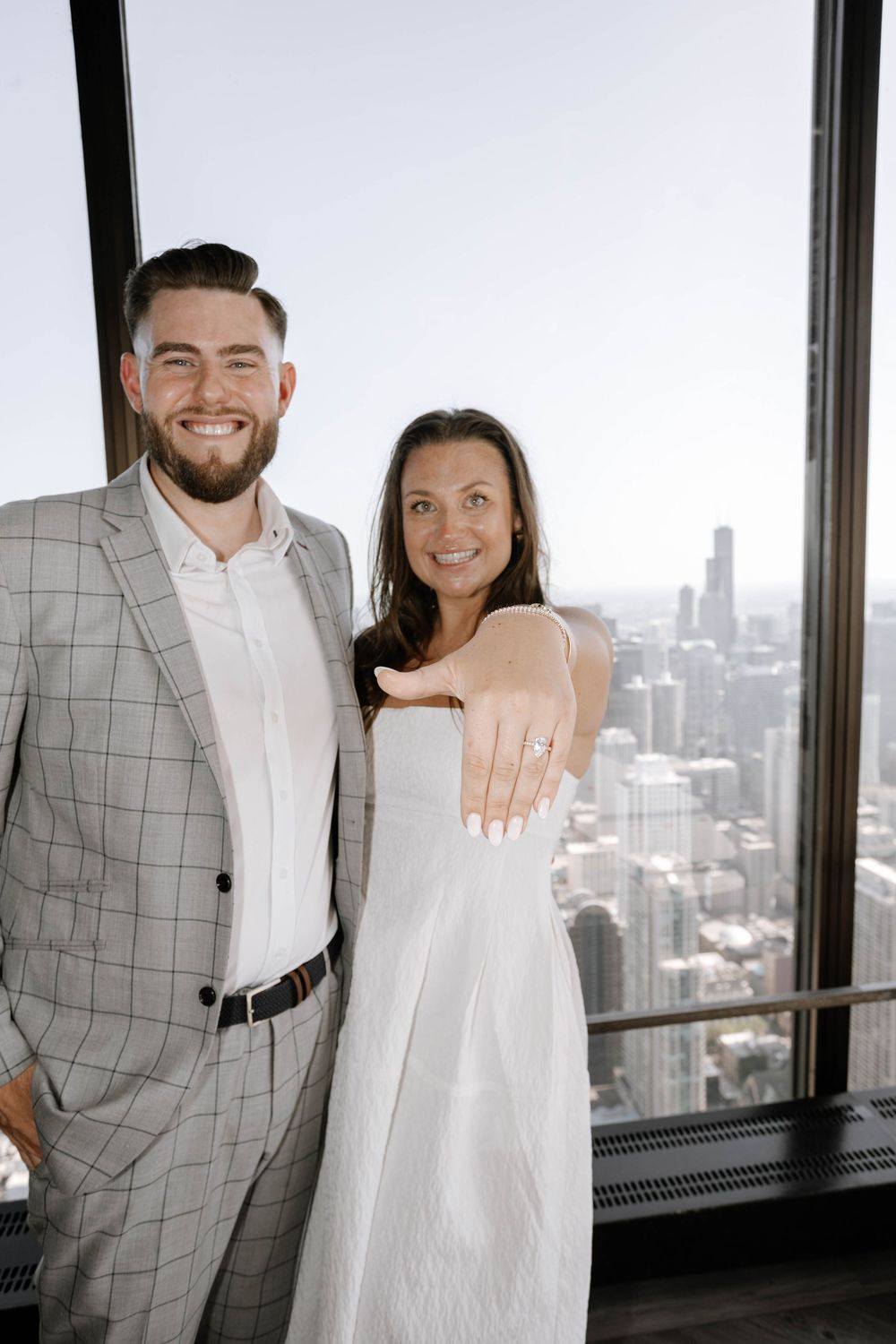 A newly engaged couple poses together in front of a Chicago skyline view from a high-rise building.