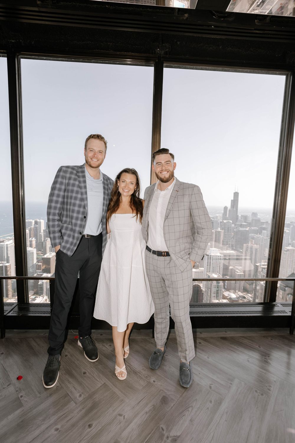 Three people pose together in front of a large window overlooking a city skyline, dressed in casual and formal attire.