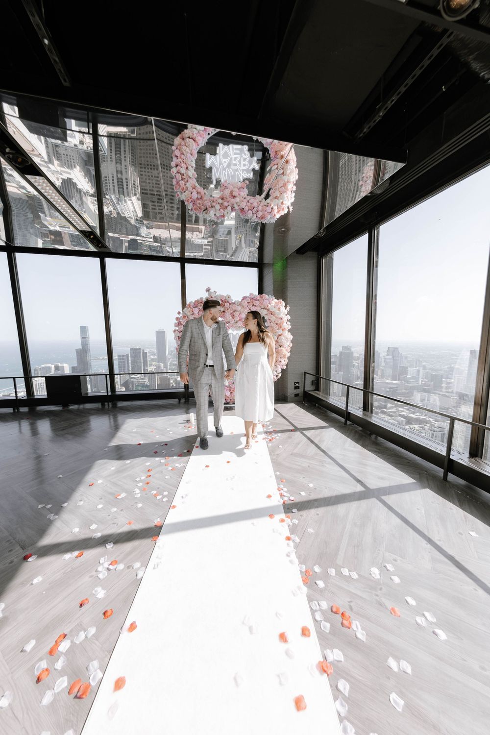 A modern high-rise event space with floor-to-ceiling windows and scattered pink rose petals creating a romantic aisle.