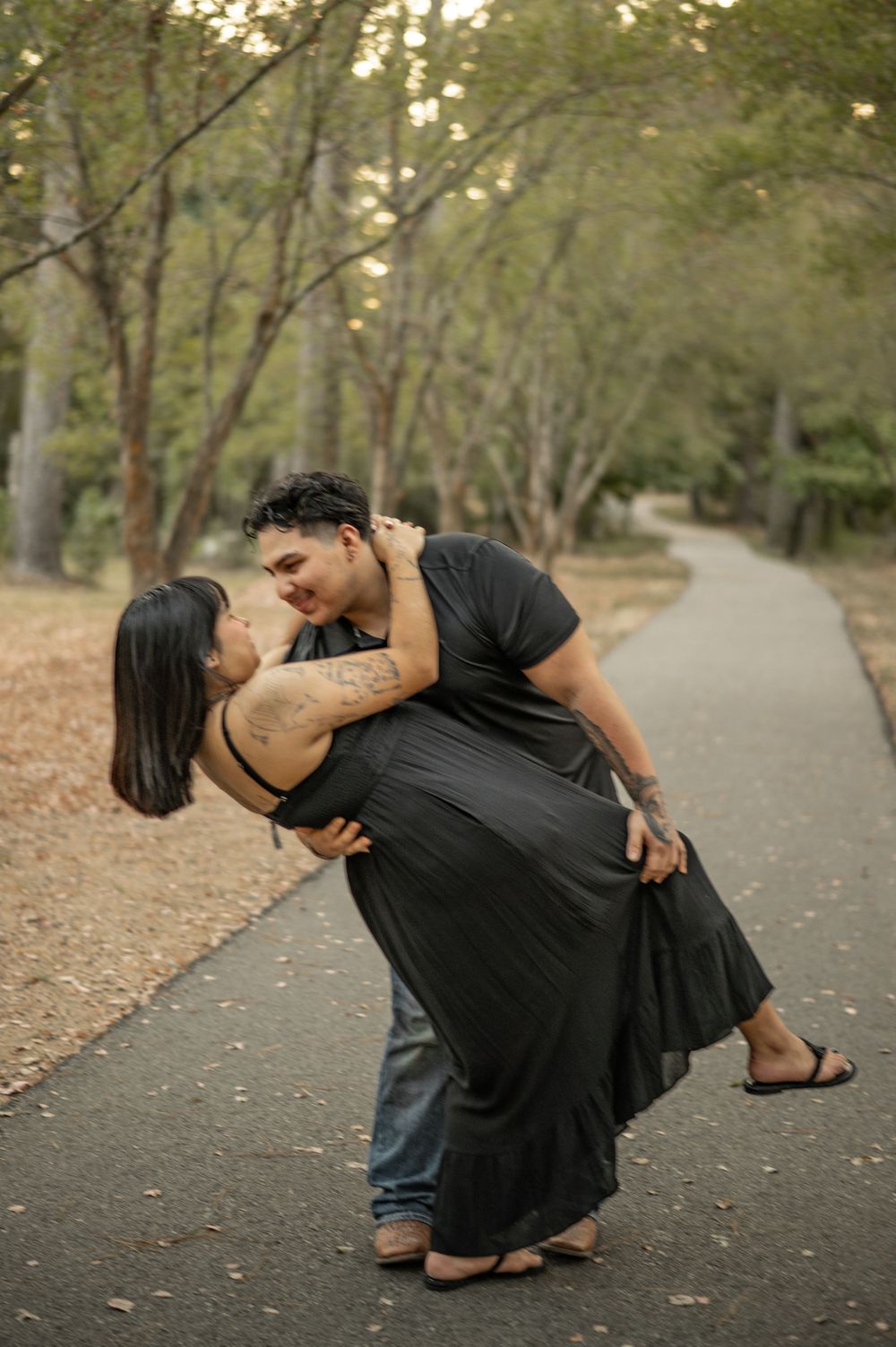A romantic couple in black outfits share an intimate dip pose on a tree-lined path during golden hour.