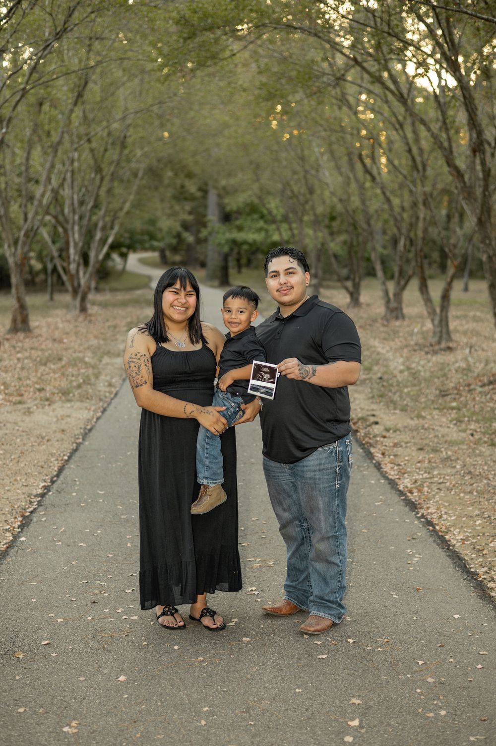 A family stands together on a tree-lined path during a casual outdoor photo shoot.