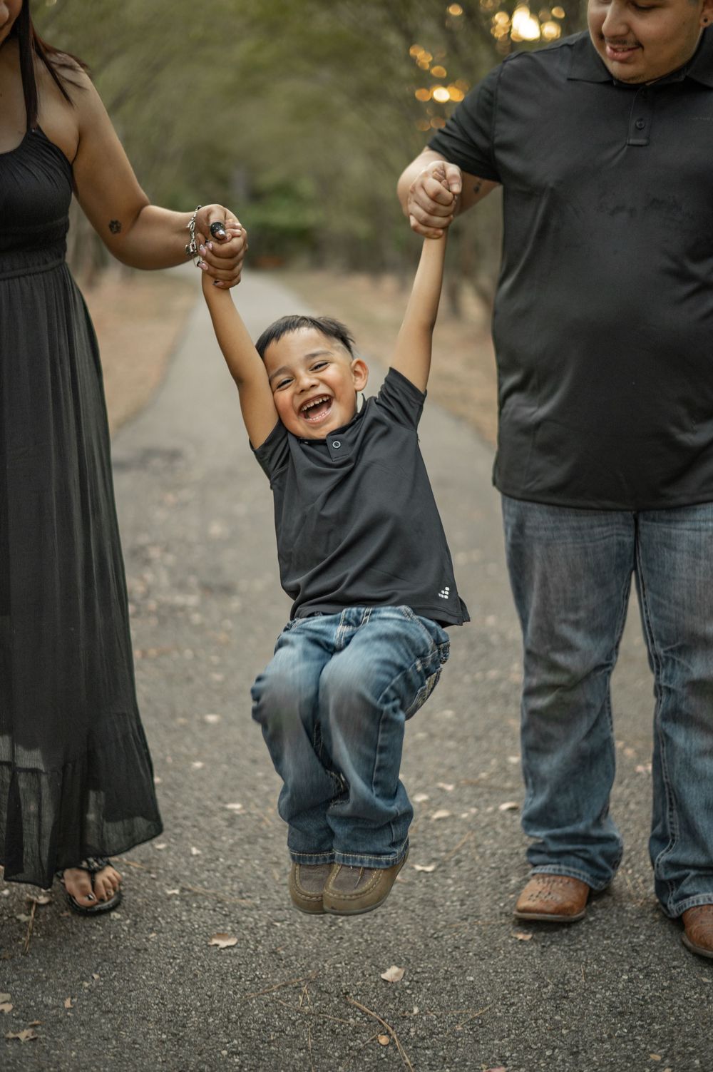 A joyful child in jeans and a dark shirt swings between two adults holding their hands on a gravel path.