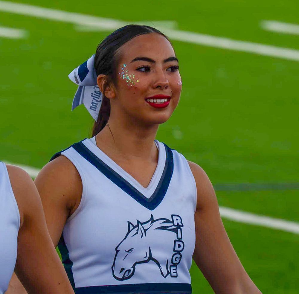 Smiling Ridge Mustangs cheerleader with glitter face paint in blue and white uniform on field.