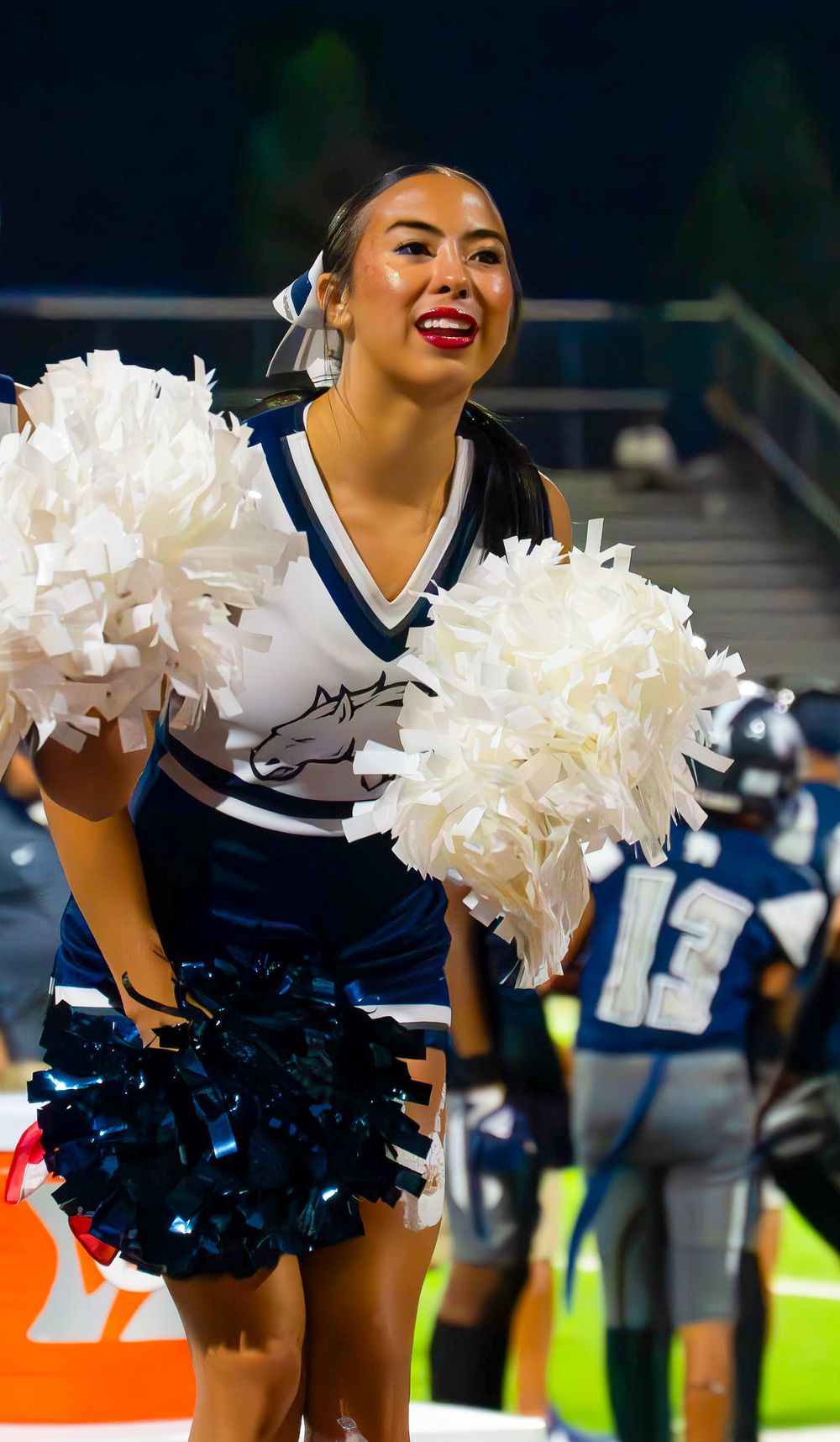 Cheerleader smiling with white and blue pom-poms in navy uniform at nighttime football game.