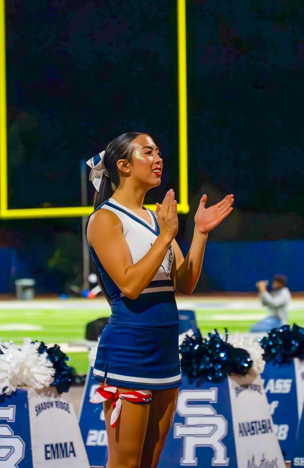 Shadow Ridge cheerleader clapping at a night football game with SR banners in background.