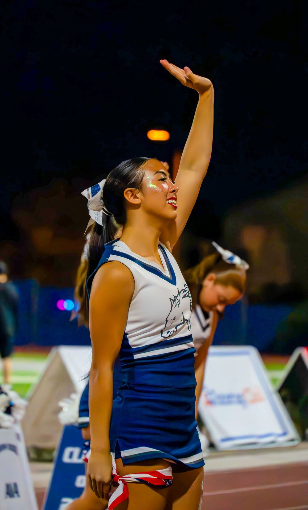 High school cheerleader raises arm performing routine in blue and white uniform at night game.