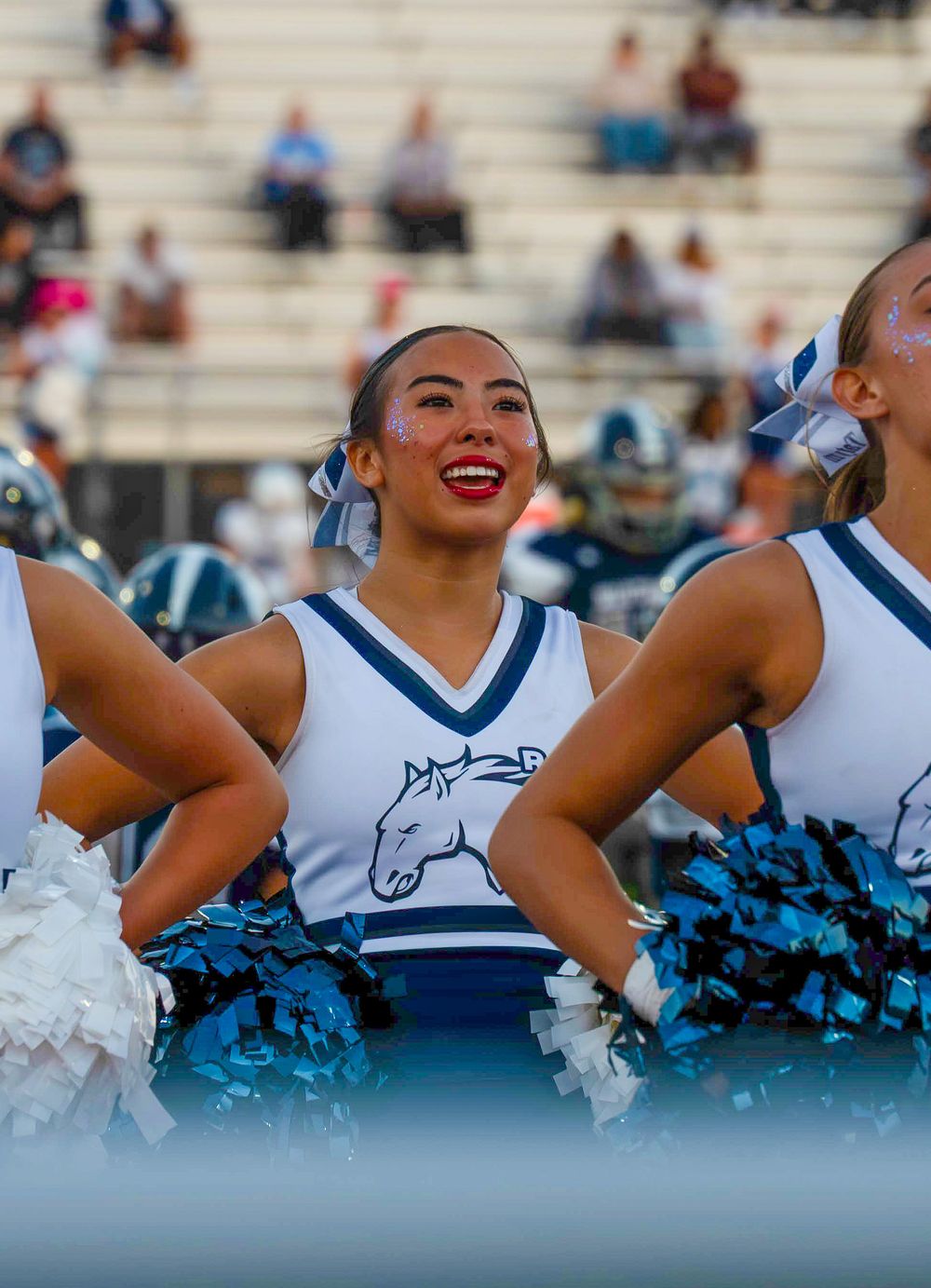 Shadow Ridge cheerleader smiling with teal pom-poms at a high school football stadium.