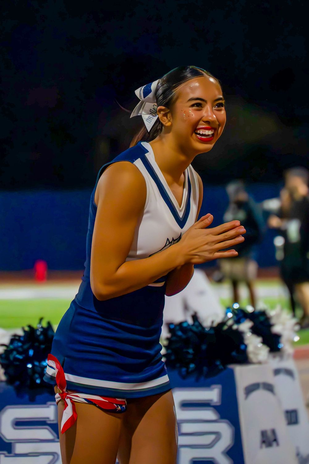 Joyful Shadow Ridge cheerleader laughs and claps in blue uniform at night football game.