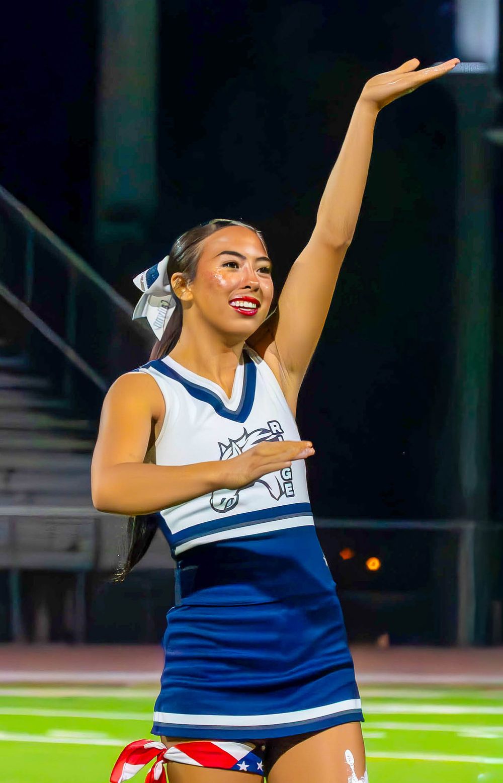 Smiling cheerleader in blue and white uniform performing routine on football field at night.