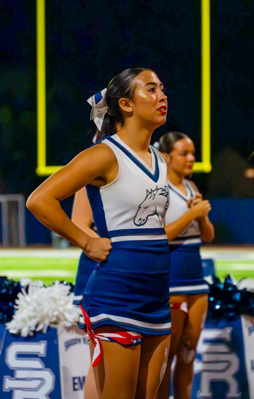Shadow Ridge Mustangs cheerleader stands confidently with pom-poms at night football game.